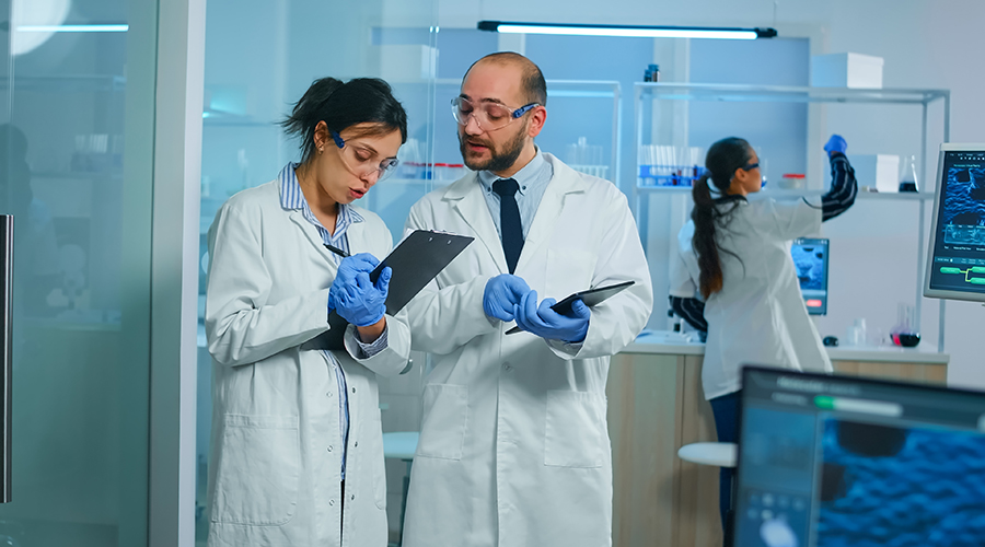 Group of medical researchers discussing about vaccine development standing in equipped lab pointing on tablet and taking notes. Doctors examining virus evolution using high tech researching diagnosis